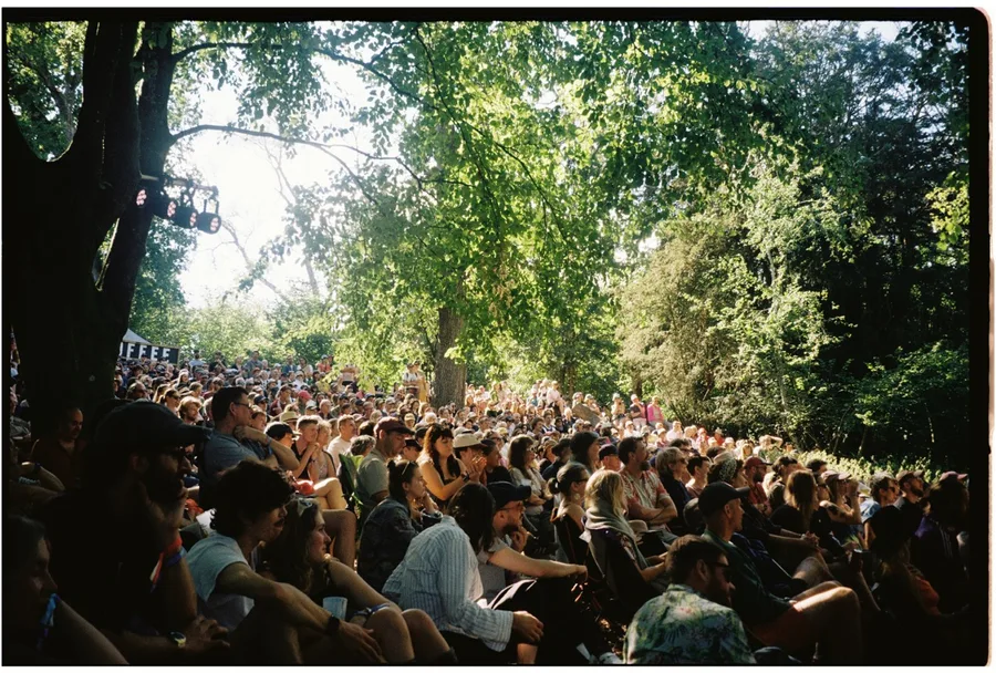 A festival crowd watching comedy from the grass under the trees